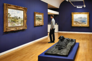 An adult with light skin and short brown hair stands in a gallery with light wooden floors and deep blue walls. Three oil paintings hang on the walls, while a bronze sculpture of a sphinx is displayed on a blue plinth.