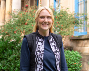 An adult with light skin, long straight blond hair, and a dark blue jacket with a white floral pattern.
