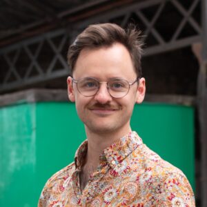 An adult with light skin, short brown hair, glasses, and a patterned yellow shirt.