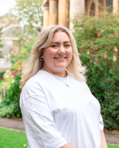 An adult with light skin, long wavy blonde hair, and a white t-shirt, standing in a garden.