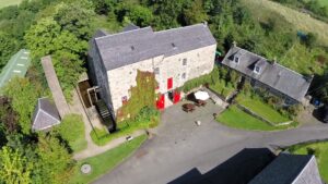 A light grey stone building consisting of 3 floors. There is bright red doors on the ground level. The building is surrounded by green trees.