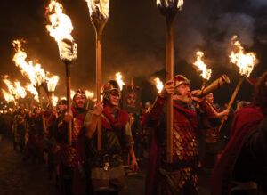 A procession of adults wearing Viking military costumes and carrying flaming torches. One of the adults is blowing a horn.