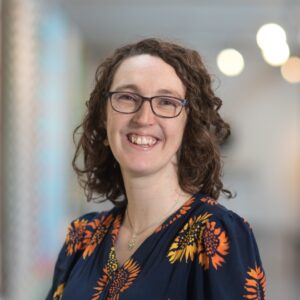 A head and shoulders photo of a person smiling with brown mid length curly hair, light skin tone, glasses.