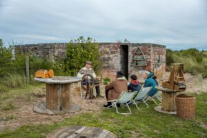An adult with light skin, short hair, and a brown leather flat cap sits at a wooden spinning wheel outside a simple brick building. A group of two adults and two children sit in folding chairs facing the spinning wheel.