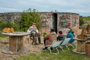 An adult with light skin, short hair, and a brown leather flat cap sits at a wooden spinning wheel outside a simple brick building. A group of two adults and two children sit in folding chairs facing the spinning wheel.
