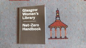 A handbook which reads "Glasgow Women's Library. Net Zero Handbook". The book is lying on a grey carpet with a red gazebo style structure.