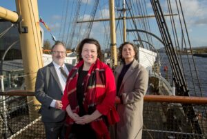 Three adults standing on board a ship. Tall masts can be seen in the background.