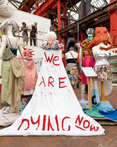 A collection of mannequins displaying outfits made of recycled materials. In the middle is a mannequin wearing a white cape which displays the words "We are dying now" in red letters.
