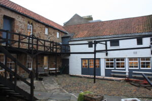 A courtyard with a cobbled ground. There is a traditional 16th-century house on the left, made from brown bricks. On the right there is a white building with a red tile roof.
