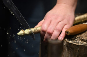 A hand with light skin holds a wooden branch. The tip of the branch has been worked into a point. A saw is cutting through the branch.