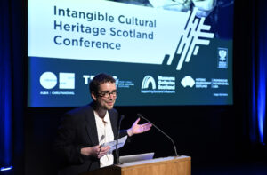 An adult with short brown hair and glasses stands at a lectern. They wear a white shirt with a smart black jacket. Behind them is a projection of a PowerPoint presentation, which reads "Intangible Cultural Heritage Scotland Conference".