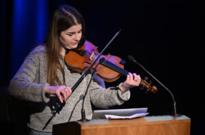 An adult with light skin, long brown hair and a grey jumper plays the violin behind a lectern.