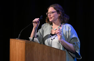 An adult with shoulder length brown hair and glasses stands at a lectern. The adult wears large necklaces and a silvery grey top.