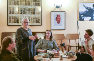 A group pf adults sit around a round table. One of the adults holds a violin. The table has several plates, cups, glasses and a teapot on it. One of the adults is standing up and addressing the rest of the group.