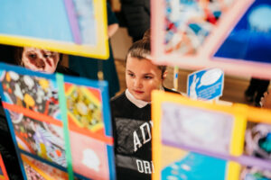 A child with light skin and dark hair in a bun looks at artwork which is suspended in the middle of the room.