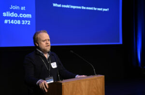 An adult with gingery grey hair and beard stands at a lectern. Behind them is the projection of a PowerPoint presentation.