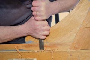 A pair of hands with light skin tone hold a chisel cutting some wood marked with pencil lines.