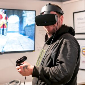 An adult in a black hoodie wears a VR headset and holds a controller whilst smiling. Behind him, a TV screen displays what the VR output.
