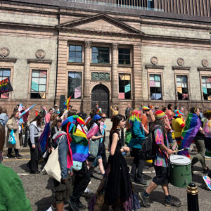 A crowd of people wearing bright colours and waving flags marches past a large stone building which features a sign reading 'Art Gallery'.