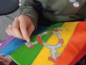 A light-skinned hand sews a transgender symbol onto rainbow fabric.