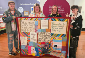 Four people stand in a gallery holding a colourful patchwork banner. There is a young person with light skin, long brown hair, and a demin jacket covered in badges; an adult with light skin and shoulder-length blonde hair; an adult with light skin, shoulder-length light brown hair and glasses; and a young person with light skin, short wavy dark brown hair, and a black jacket covered in studs.