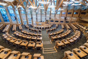 An ariel view of the debating chamber of the Scottish Parliament. Rows of black chairs at wooden desks face a large wooden podium.