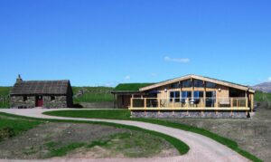 A path leading up to two buildings. The building on the left is a small cottage with grey stone walls and a thatched roof. The building on the right has a wood panelled front with a large decking area outside. The roof is covered in green grass.
