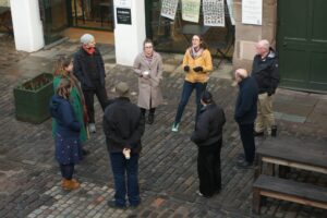 A group of adults wearing winter coats stand in a circle in a courtyard with flagstone flooring.