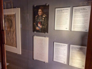 A display case featuring four labels, a handwritten letter, and a portrait of an adult with light skin and an elaborate blue gown.