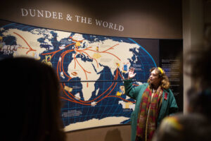 An adult points to a map of the world, focused on Dundee. Above the map, some text reads 'Dundee & the World'.