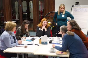A group of adults sit at a table and engage in conversation. Behind them is an adult standing next to a flipchart.