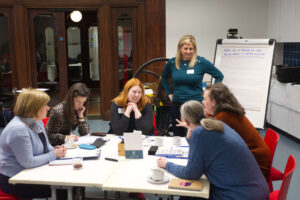 A group of adults sit at a table and engage in conversation. Behind them is an adult standing next to a flipchart.