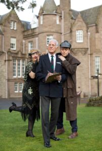 Three people with light skin tone stood together. In the middle the person with short grey hair in a suit is holding a clipboard whilst the others wearing 1920s historical costumes look on.