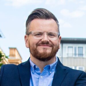 An adult with light skin tone, short grey hair, and a red beard. They are wearing a pair of glasses, a blue shirt, and a blue blazer.