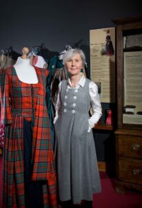 A person with light skin tone and light grey short hair is stood next to a mannequin wearing a red and blue long tartan dress. In the background are panels of text and historical costumes on hangers.