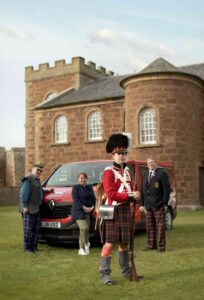 Three people with light skin tone are stood in front of a red minivan parked on a lawn in front of a stone building. In the foreground a person with light skin tone wearing a kilt, red soldier's uniform jacket, and tall black hat is holding a rifle.