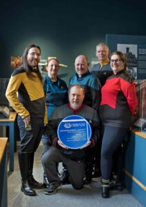 6 people with light skin tone stood together wearing matching tops in yellow, blue, red, and grey. In the middle a person is kneeling holding a blue plaque.