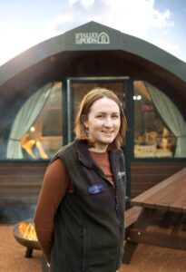 A person with light skin tone stands in front of a fire pit, picnic bench, and domed small wooden building.