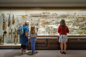 An adult and two young people with light skin stand in front of a display of brass instruments.