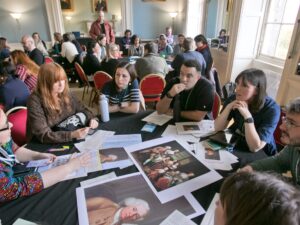 Groups of adults sit around circular tables and engage in discussion. Print-outs of 18th century portraits are spread across the tables.