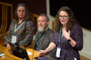 An adult with light skin and long wavy brown hair stands and speaks at a podium.
