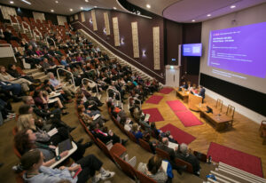 An adult with light skin and short black hair addressing an auditorium full of people.