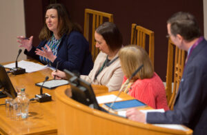 An adult with light skin and shoulder-length brown hair speaks into a microphone as three adults listen.