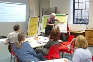 Five adults sit at tables and look at one person, standing and presenting to the group
