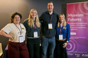 Four adults of different heights stand together smiling. They have light skin, one has curly brown hair pulled back, two have medium length blonde hair and one has very short brown hair. Behind them, a pink and purple pull-up banner with the title 'Museum Futures'