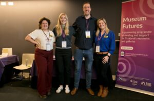 Four adults of different heights stand together smiling. They have light skin, one has curly brown hair pulled back, two have medium length blonde hair and one has very short brown hair. Behind them, a pink and purple pull-up banner with the title 'Museum Futures'