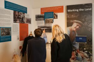Two adults, one with short brown and hair and one with medium length blonde hair, stand facing away frm us pointing and looking at museum display panels on a wall.