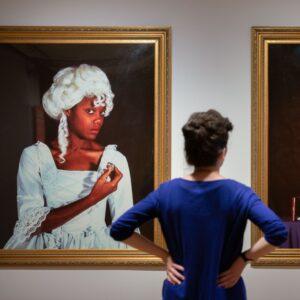 An adult with light skin and brown hair tied into a bun views a portrait by Maud Sulter, a Scottish artist of Ghanaian heritage, at Edinburgh’s City Art Centre. The portrait depicts an adult with medium-dark skin wearing a white wig and formal dress in the style of 18th century Western fashion.