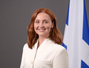 An adult with light skin tone and shoulder length red hair. They are wearing a white suit jacket. A saltire is on display behind them.