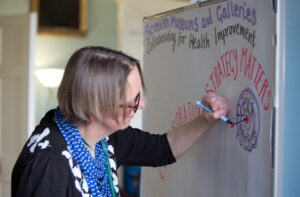 An adult with light skin tone and short grey hair. They are drawing on a large piece of paper stuck to a whiteboard.